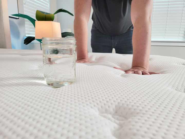 An image of a glass of water on top of the Titan Plus Luxe, with a man pressing into the mattress around the glass.