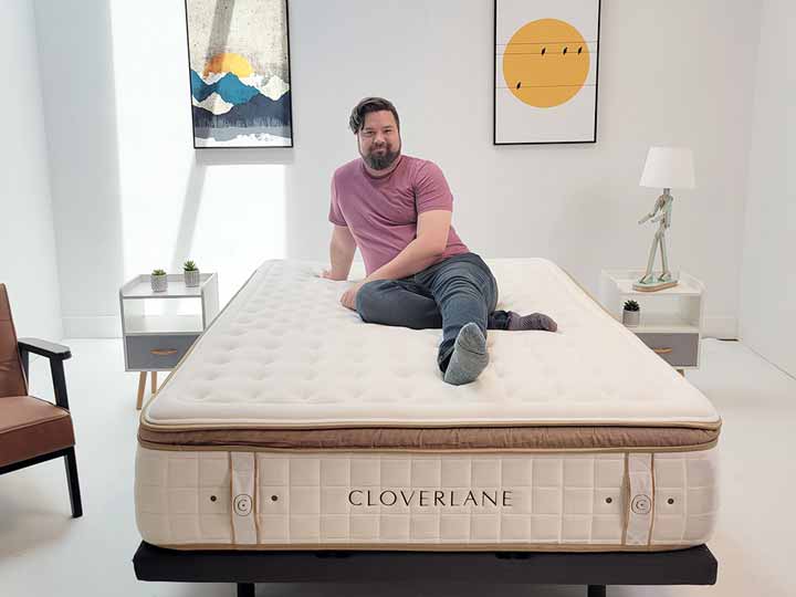 An image of a man sitting on top of the Cloverlane Hybrid Mattress, smiling at the camera.