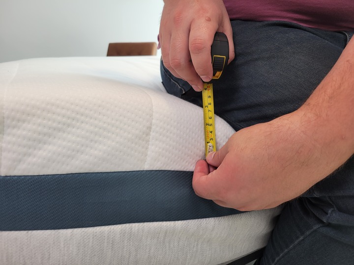 A man sits on the edge of the Helix Twilight mattress with a tape measure displaying about 3 inches of sagging